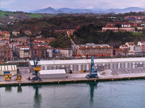 Pasai Donibane, Spain - 10 March 2026: Aerial view of cranes standing tall against the backdrop of the harbor and quaint buildings, reflecting in the calm water.