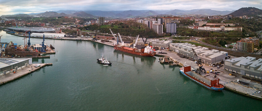 Pasai Donibane, Spain - 10 March 2026: Aerial view of the harbor where ships rest between industrial docks beneath a skyline of buildings and rolling green hills.