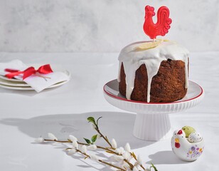 A Russian-style chocolate Easter cake decorated with icing, white chocolate feathers, and a red caramel cockerel on a white stand against a light background. © Natallya_ph