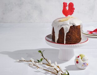 A Russian-style chocolate Easter cake decorated with icing, white chocolate feathers, and a red caramel cockerel on a white stand against a light background. © Natallya_ph