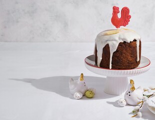 A Russian-style chocolate Easter cake decorated with icing, white chocolate feathers, and a red caramel cockerel on a white stand against a light background. © Natallya_ph