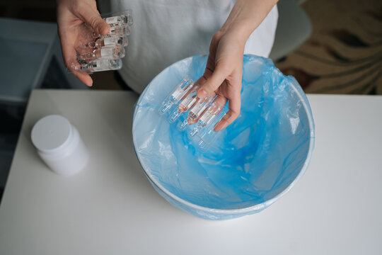 Top view of person hands dropping multiple glass ampoules of expired medication into blue plastic garbage bag inside white trash can, illustrating proper and safe disposal of pharmaceutical waste