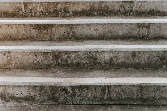 A close-up of worn concrete stairs with visible stains, cracks, and rough texture forming a repeating pattern throughout the frame. The neutral tones create a minimalist urban backdrop.