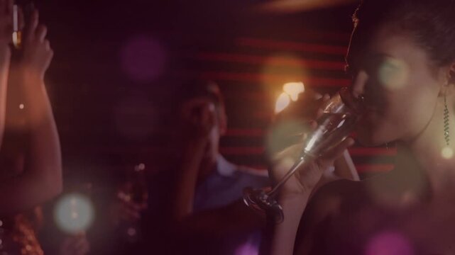 Woman sipping in dim party lounge, camera pulling back showing group raising flutes toasting, bokeh