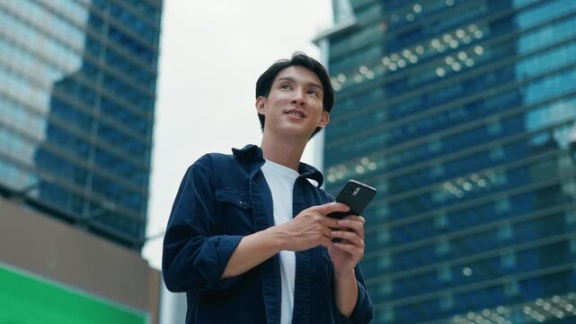 Portrait of young Asian man standing outdoor happy smiling use mobilephone.Handsome young asian man dressed casually walking spending time outdoors at city urban,carrying backpack,using mobile phone
