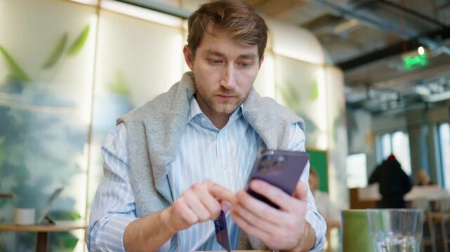 Caucasian man holding credit card and smartphone while checking payment details at cafe table. Adult male comparing banking information and mobile screen before online purchase. Digital payment moment