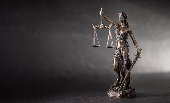 A bronze statue of Lady Justice on a dark, textured background with strong side lighting that highlights her details and symbolism. The composition, with free space on the left