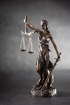 A bronze statue of Justice stands against a dark, textured background with dramatic side lighting. The figure holds scales symbolizing justice in one hand and a sword in the other,