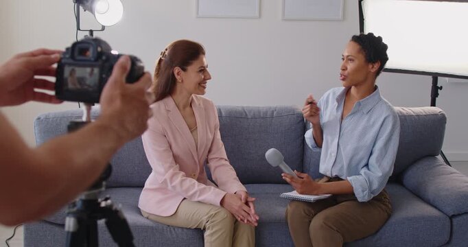 Studio interview recording, meeting and conversation of journalist with microphone and female guest sitting on sofa. Professional cameraman filming video, giving thumbs up to reporter talking to woman