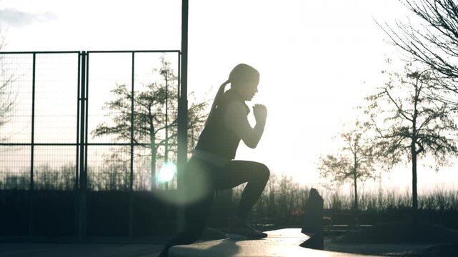 Silhouetted figure in sportswear lunges in sunlit outdoor space, bordered by a chain-link fence and sparse trees. Moment of focused sport training, blending athleticism with serene nature backdrop.