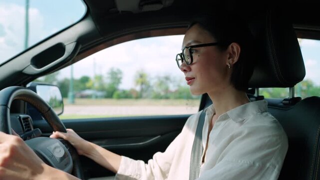 Happy Asian woman wearing eyeglasses driving a car and wearing a seatbelt. Smiling female driver enjoying a road trip or daily commute. Concept of safe driving, auto insurance, and travel.