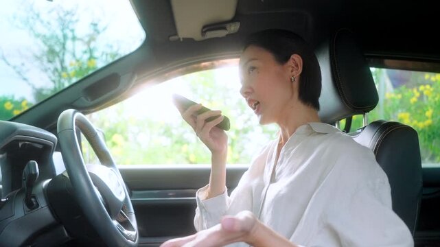Frustrated Asian woman sitting in the driver's seat of her car, intensely talking on her smartphone using speakerphone, gesturing with her hand in annoyance over a bad phone call.