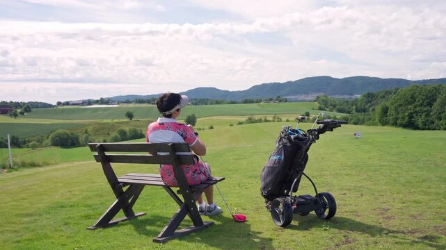 Senior woman sitting on a wooden bench, resting during a golf game with a scenic mountain backdrop. Authentic mental health break, sustainable wellness, and peaceful retirement lifestyle concept.