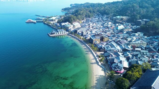 Drone aerial landscape of Mikasa Beach ocean shoreline with suburban town housing streets and village shops with ferry port pier in background of Miyajima Island Hiroshima Japan travel and tourism