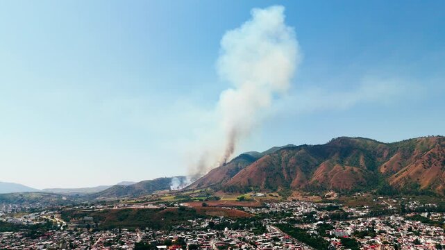 Dangerous forest fire burning on a mountain, sending a large column of smoke over a city in mexico