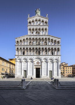 View of the Church of San Michele in Foro, a symphony of white marble tiers rising against a clear blue sky, casting long shadows on the cobblestone square, Lucca, Tuscany, Italy.