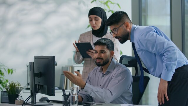 Arabian business man working with computer in office two colleagues come in Muslim businesswoman secretary with Indian boss leader check work online helping looking at screen three co-workers talking