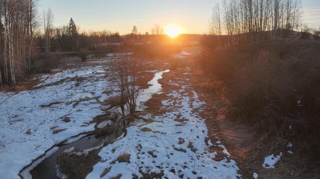 Aerial view of a partially frozen creek meandering through a marshy landscape with snow patches at sunset near Whitefish, Montana.