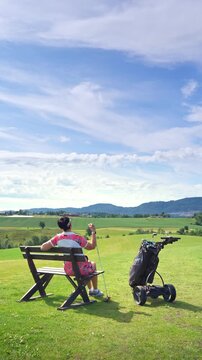 Senior woman sitting on a wooden bench, resting during a golf game with a scenic mountain backdrop. Authentic mental health break, sustainable wellness, and peaceful retirement lifestyle concept.