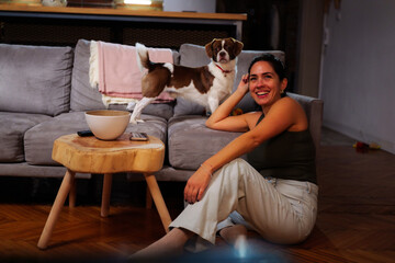 Woman sitting on the floor smiling while her small dog stands on the couch behind her, sharing a...
