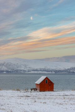 View of a rustic red cabin, dusted with snow, stands sentinel against the backdrop of serene waters and snow-capped mountains, under a crescent moon, Lyngen, Norway.