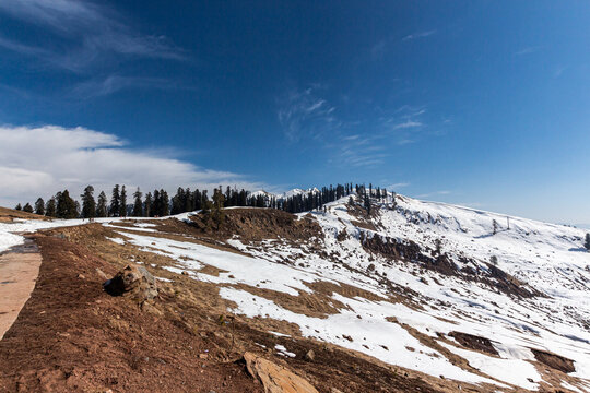 View of snow-covered mountain slopes meet bare earth under a bright blue sky dotted with wispy clouds, a contrast of winter's touch and nature's resilience, Bagh, Azad Kashmir, Pakistan.