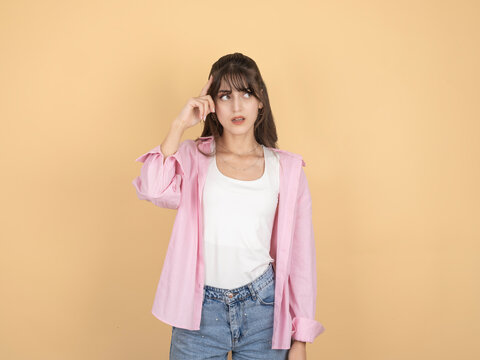 Thoughtful young woman touching her temple and looking aside, wearing pink shirt and denim, standing against beige studio background with copy space. Thinking and decision concept.