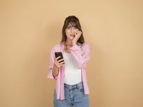 Nervous young woman biting her nails while holding smartphone, wearing pink shirt and denim, standing against beige studio background with wide copy space. Anxiety and stress concept.