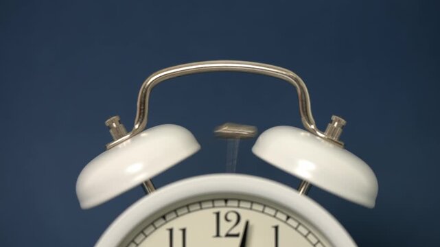 A classic white mechanical alarm clock vigorously rings, its small hammer rapidly striking the twin bells atop a dark blue background.