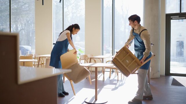 Cafe workers carrying chairs and talking in restaurant hall. Man and woman arranging furniture and smiling at each other. Teamwork concept. Staff organizing dining area together in spacious large cafe