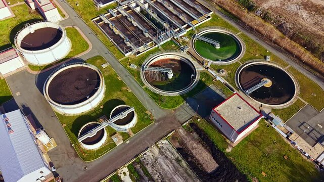 A water treatment facility works on purifying wastewater in various large circular tanks during daylight. The location features multiple structures and greenery surrounding the area.