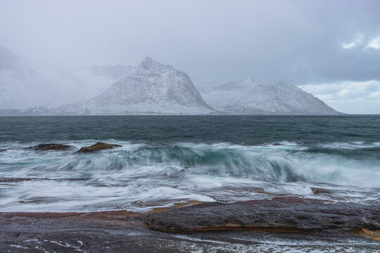 View of icy waves crash against the rocky shore, the distant snow-covered mountains loom under a gray sky, creating a stark contrast of textures, Senjahopen, Troms, Norway.