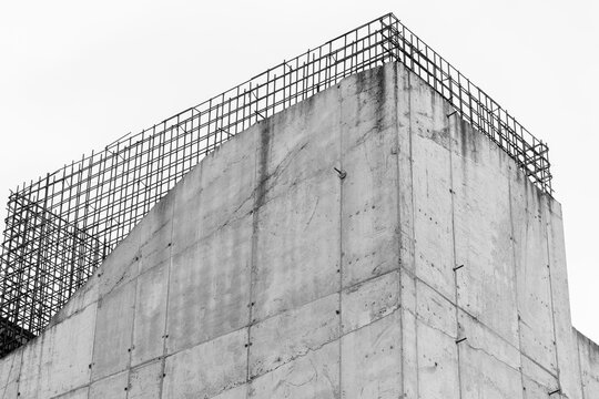 A stark black-and-white photograph of a concrete building corner with exposed rebar grid, highlighting industrial textures, geometric lines and minimalist architectural detail in an urban construction