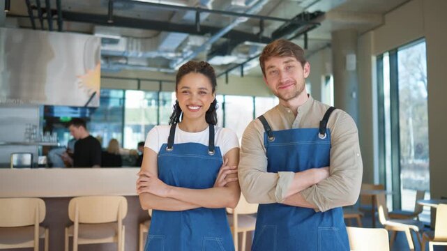Cafe workers standing with crossed arms in restaurant interior. Man and woman wearing aprons smiling at camera. Staff members posing together near bar counter inside modern dining hall.