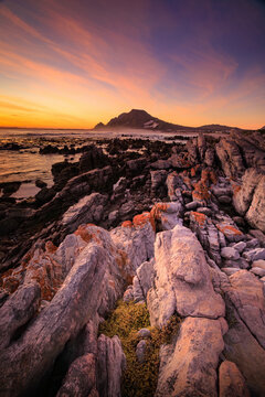 View of rugged, textured rocks lead the eye towards the distant mountain under a vibrant sunset sky, Betty's Bay, Western Cape, South Africa.