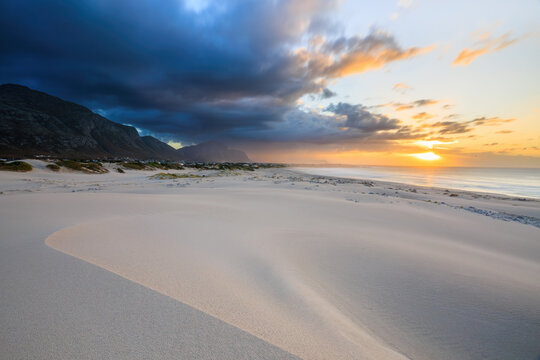 View of a serene sandy beach meeting the ocean under a dramatic sky with dark clouds contrasting with the warm glow of the setting sun, Betty's Bay, South Africa.
