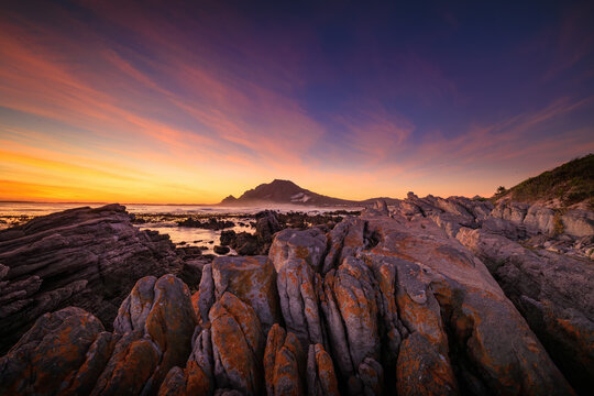 View of rugged, lichen-streaked coastal rocks meet a fiery sunset sky over the distant mountain silhouette, Betty's Bay, Western Cape, South Africa.