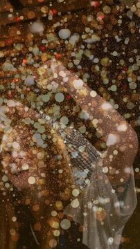 Vertical video: Woman celebrating in bar arm lifting sparking gold confetti over cap warm lights