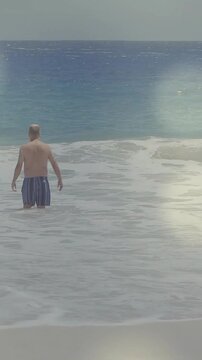 Vertical video: Retiree entering shallow ocean, seeking open water with breaking waves for travel
