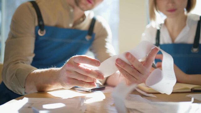 Close view of baristas checking receipts and comparing payment data. Hands holding paper bills and reviewing numbers together. Writing notes and organizing documents on table. Managing finances.