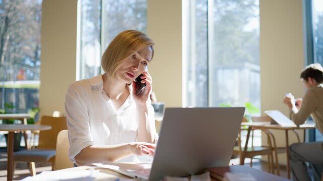 Pretty Caucasian female sitting at table and talking on smartphone. Looking down at documents and checking details. Holding phone near ear and focusing. Laptop nearby. Remote communication concept.