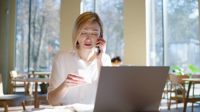 Charming woman sitting at table with laptop and phone. Holding receipt and talking on call. Asking advice and discussing issue. Looking focused and serious. Papers on desk. Indoor cafe.