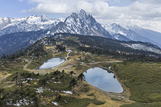 Aerial view of twin lakes nestled amidst the rugged terrain of Peacock Mountain, with snow-capped peaks piercing the skyline, Deqing, Yunnan, China.