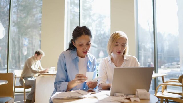 Two women sitting at table with laptop. One holding receipts and reading details. Other typing and focusing on screen. Checking expenses and records together. Papers scattered on desk. Cafe interior.