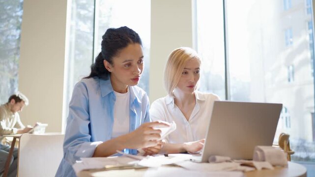 Two women sitting at table and working with laptop. One sorting receipts and reading numbers. Other typing and checking data on screen. Discussing financial details together. Papers scattered on desk.