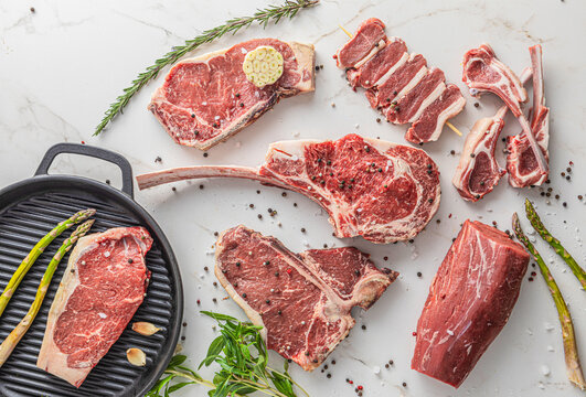 Top view of various premium raw beef cuts including Tomahawk, T-Bone, ribeye, and lamb chops on white marble, with a cast iron grill pan, asparagus, and spices, concept for butcher shop and gourmet co