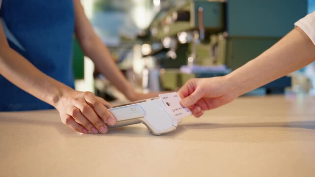 Client holding card near payment terminal at cafe counter. Barista preparing device for contactless transaction. Making purchase using bank card at checkout. Close view of modern payment process.