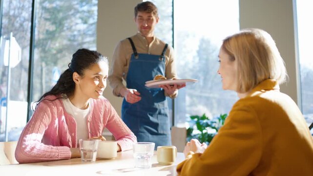 Male barista approaching table with tray and breakfast order. Female customers looking at worker while waiting for food. Holding plates and preparing to serve meal. Interaction happening inside cafe.