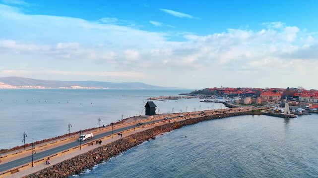 Wide aerial shot of a causeway with a wooden windmill. Scenic view of a road over the sea featuring a traditional dark wooden windmill landmark on the side.
