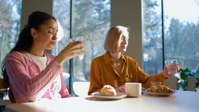 Friends drinking water while sitting at cafe table. Woman lifting glass and taking sip during meal. Friend holding drink and pausing during conversation. Eating croissants and resting midday break.
